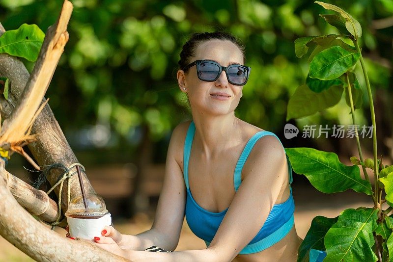 美丽的白人女性戴着太阳镜和蓝色比基尼，在海滩旁郁郁葱葱的热带树木公园里喝着混合冰饮料