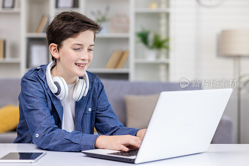 快乐的十几岁男孩，脖子上戴着耳机，坐在明亮的房间里的笔记本电脑前，敲击键盘，玩游戏，监控社交网络，做作业