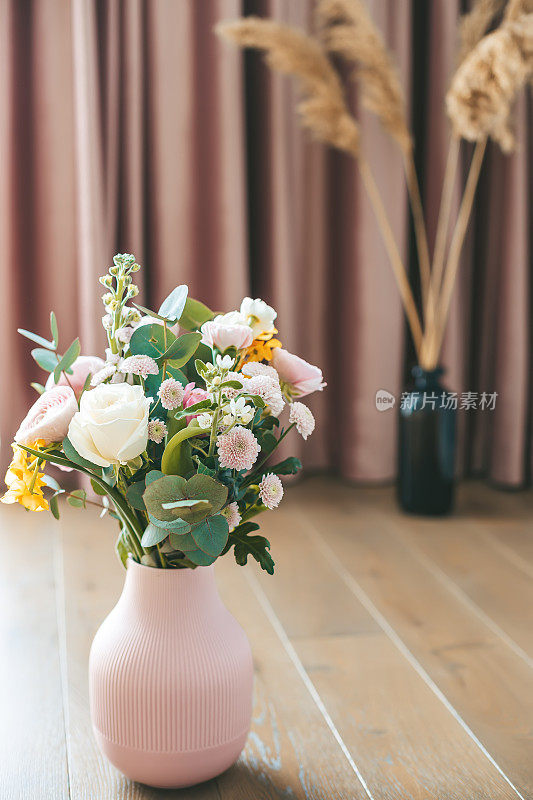 在一个有纹理的粉色花瓶中，各种各样的花朵与优雅的粉色窗帘和黑色花瓶中的干燥植物相衬