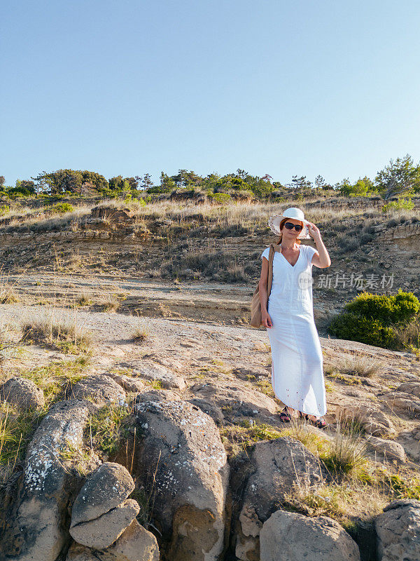美丽的女人，穿着白色的夏装，戴着太阳帽，站在岩石嶙峋的海边
