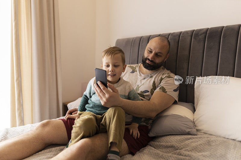 爸爸和小儿子笑着看着手机上的照片。他们坐在卧室的床上拍照。父亲拥抱儿子。他们做鬼脸。一个黑发浓密的男人和一个金发碧眼的男孩。焊接时间