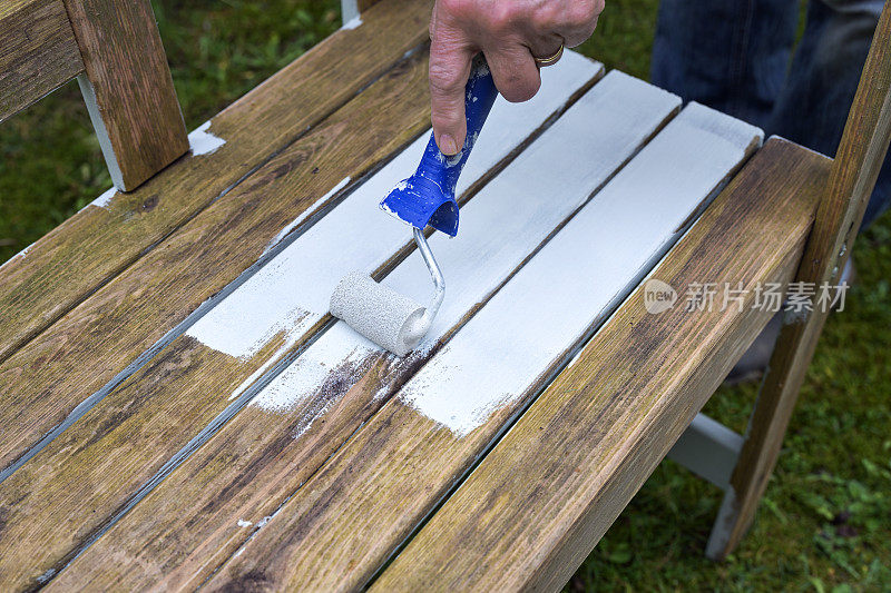 用油漆滚筒在风化的花园木凳上涂上白色的扩散开放式天气防护涂层，为户外季节、复制空间做准备