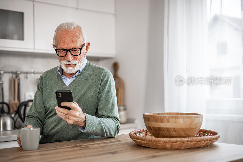 老人，退休老人，坐在厨房里，带着快乐的微笑，喝着热咖啡或茶，用着手机，早晨的例行公事，无忧无虑的退休日子