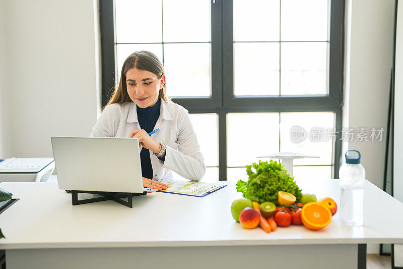 年轻的金发女营养学家拿着水果站在她面前，在办公室的办公桌前制定饮食计划