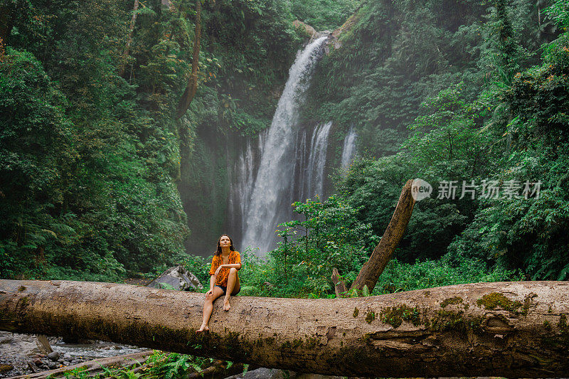 徒步旅行时，女人坐在热带瀑布背景的大原木上