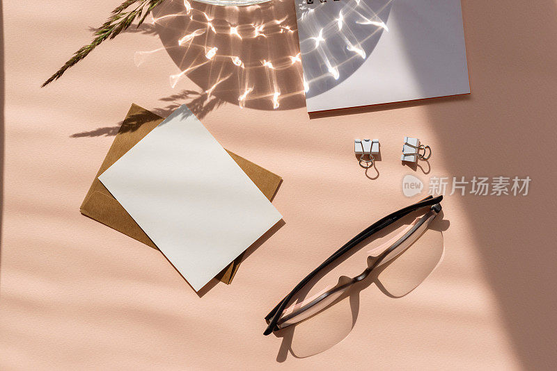 唯美女性化的小商业品牌工作空间，空白纸卡、信封、眼镜、文具在桃红色柔色背景上配以抽象自然的阳光阴影，平铺