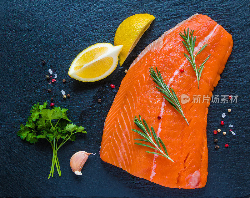 大块生鲑鱼，新鲜柠檬，迷迭香和欧芹，海盐和胡椒粉放在黑石板上，俯视图。