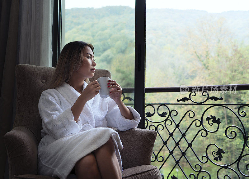 在一家豪华五星级酒店里，裹着毛巾的女人透过窗户看外面，喝着咖啡