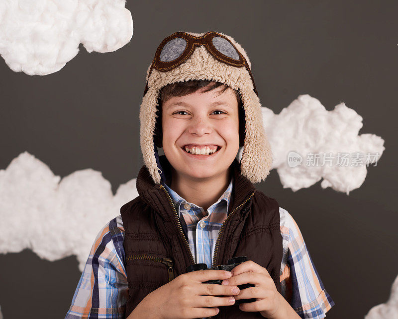 一个男孩玩着双筒望远镜，梦想着成为一名飞行员，穿着复古风格的夹克，戴着戴眼镜的头盔，棉絮缭绕，灰色的背景，略带棕色