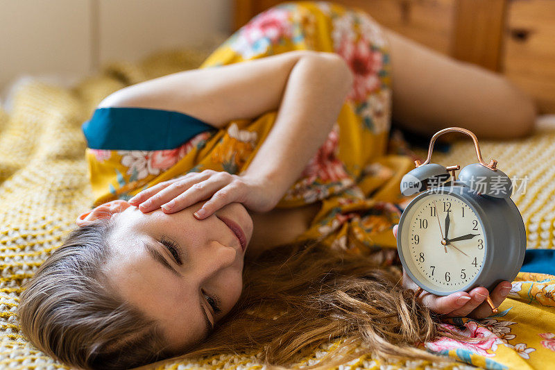 一个焦虑的金发年轻女人在床上，在早上看闹钟的时候睡过头了