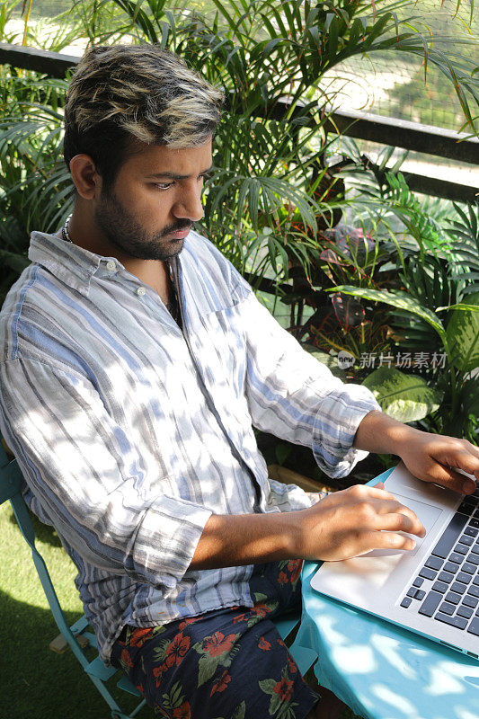 一名男子坐在阳台露台上的蓝色圆桌旁，在阳光下用笔记本电脑工作，背景是热带棕榈树丛林花园，俯瞰公寓塔楼，冠状病毒新常态在家工作概念图片集。