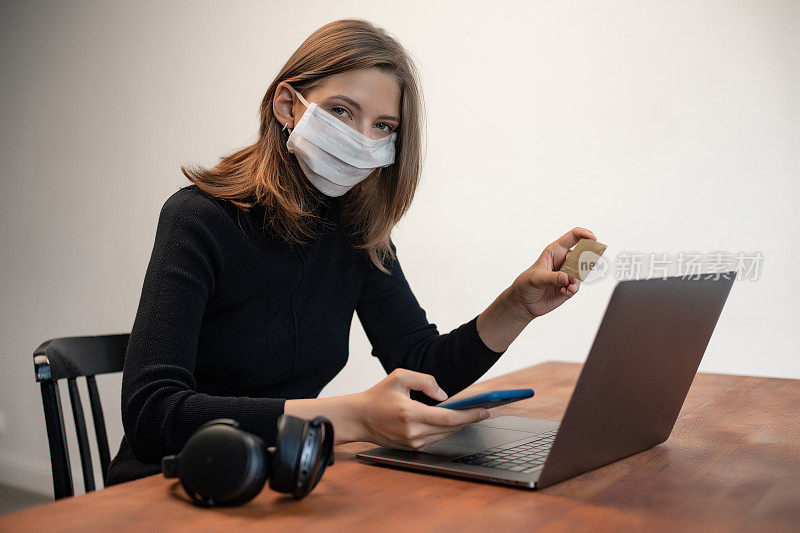 可爱的白人女孩在室内戴医用防护口罩的肖像。女人是持卡人，用塑料银行卡在笔记本电脑上上网工作