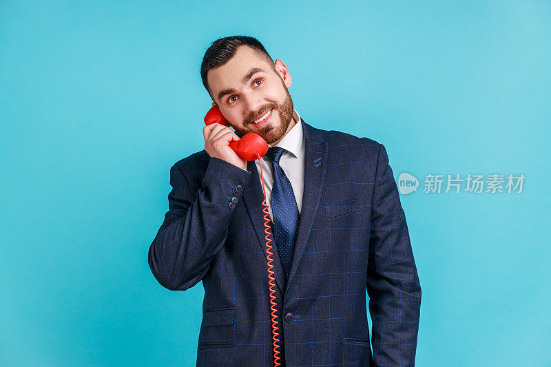 快乐的男人胡子穿着官方风格的西装谈论座机电话手持听筒，带着牙齿的微笑看着别处。