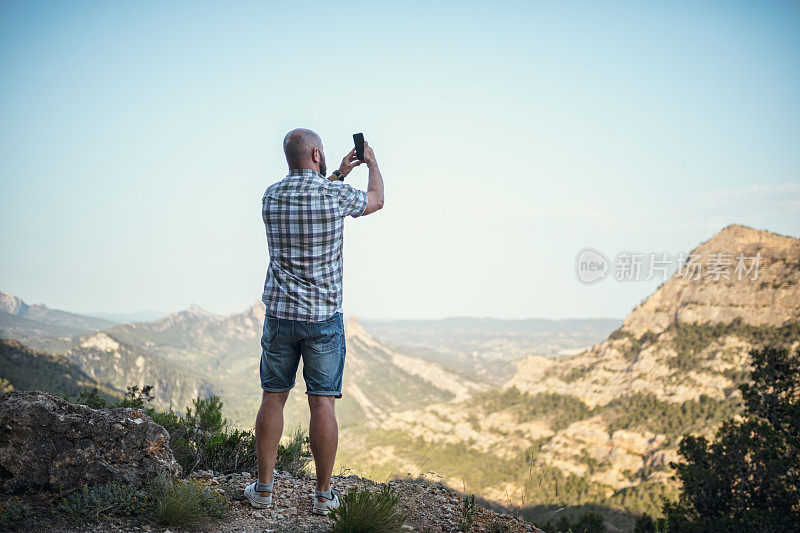 一个在山里拿着智能手机的秃顶成年男子的后视图