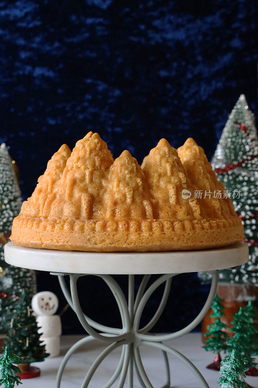 自制的图片，圣诞树形状，白色的柠檬毛绒绒的Bundt蛋糕，大理石和金属蛋糕架周围环绕着圣诞树模型，柠檬味的环状海绵覆盖着糖霜，棉花糖雪人，蓝色背景