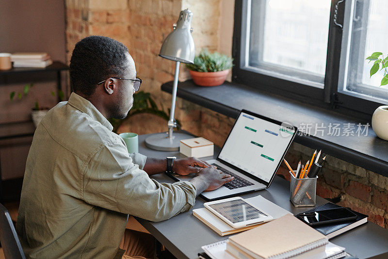 水平中景拍摄的是一名黑人坐在办公室的办公桌前，用笔记本电脑研究财务策略