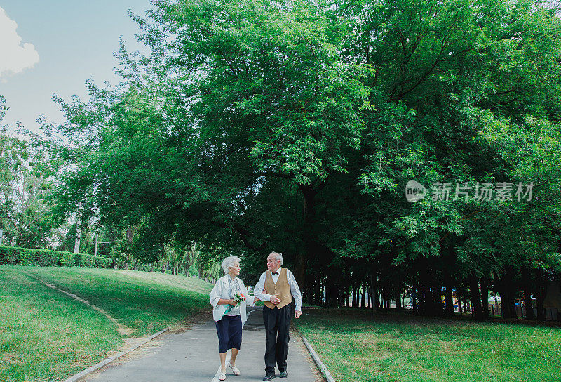 一对老夫妇在绿色的公园里散步。祖母和祖父在他们的金婚纪念日上。50年老人在一起的爱情故事。奶奶和爷爷在说话。