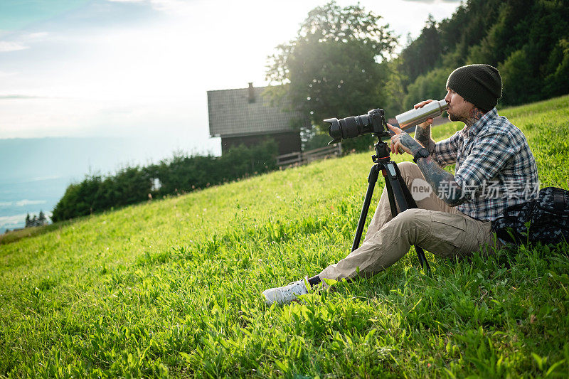 风景摄影师坐在草地上，用无反光镜相机拍摄大自然，用保温瓶喝着水。