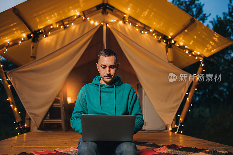 快乐的人自由职业者使用笔记本电脑在一个舒适的野营帐篷在夏夜。豪华户外露营帐篷，适合夏季度假和度假。生活方式的概念
