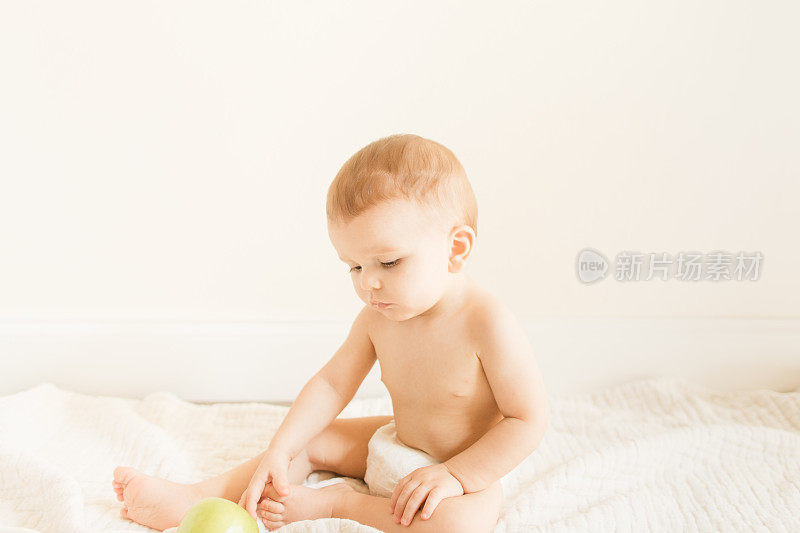 2022年秋天，一个9个月大的大流行男婴穿着可持续制作的棕色针织紧身衣，在一个新鲜的绿色史密斯奶奶苹果上玩耍和长牙，坐在家里地板上的奶油色薄纱毯子上