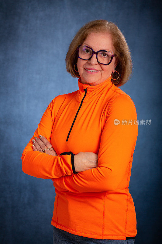 一个60多岁的女人的肖像。她站在那里看着镜头，她很生气。她穿着一件亮橙色的运动上衣，戴着一副眼镜。