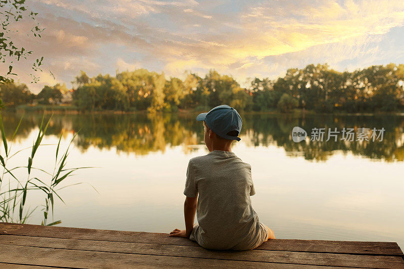 在湖边码头上的男孩。孩子看湖，看日落，看大自然。孩子坐在码头上。摄影。风景