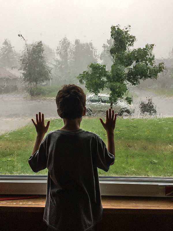 小男孩从后面透过客厅的窗户看着暴风雨