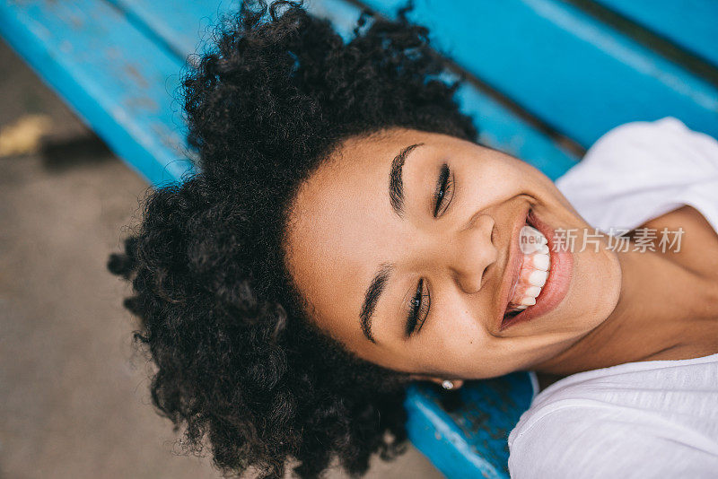 快乐的非裔美国妇女在公园的长凳上休息的特写镜头，笑着露出牙齿的微笑。美丽年轻的黑皮肤女性享受健康的生活。人与生活理念