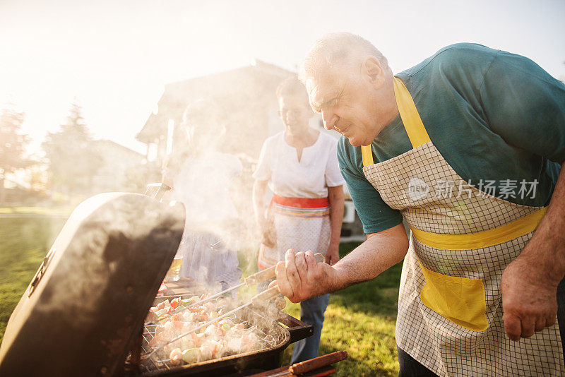 快乐的老人正在检查他的肉在烧烤，而他的家人正在享受在后院