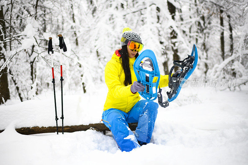 在一片冰冻的森林里，一个雪上徒步旅行者正在休息，坐下来清理雪地鞋上多余的雪