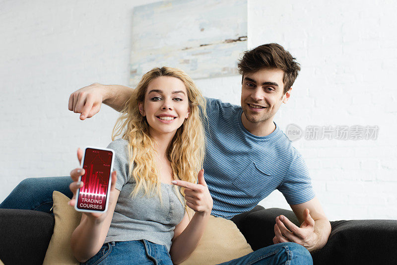 一个快乐的男人和一个金发女人用手指指着智能手机，屏幕上有交易课程的应用程序