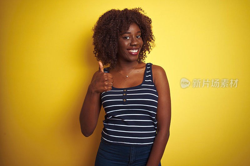 年轻的非洲黑人妇女穿着条纹t恤在孤立的黄色背景做快乐的拇指手势与手。赞许的表情看着镜头，表示成功。