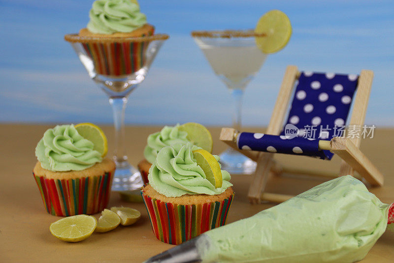马提尼杯包含自制的热带鸡尾酒，菠萝朗姆酒纸盒纸杯蛋糕，上面有绿色奶油糖霜，鸡尾酒伞和柠檬片，躺椅，沙滩和蓝天背景，聚焦前景