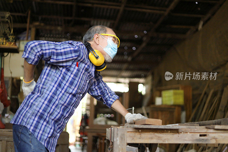 亚洲一名资深木匠在退休后的业余爱好中，在他自己的车库风格的车间里锯木板，饱受背痛之苦