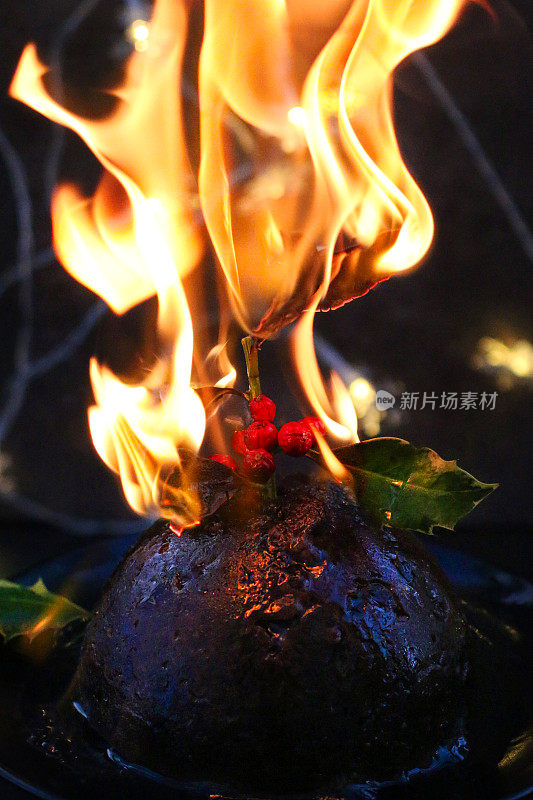 特写图像的圣诞布丁浸泡在白兰地和点燃，燃烧的橙色火焰，五颜六色的冬青树叶子与红色浆果，照明的童话灯，黑色背景，聚焦前景