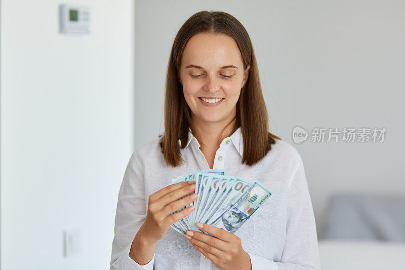 室内拍摄的非常开心的女性穿着白衬衫，手里拿着扇子的钞票，梦想着买很多东西，中了彩票，为有一大笔钱而欣喜。