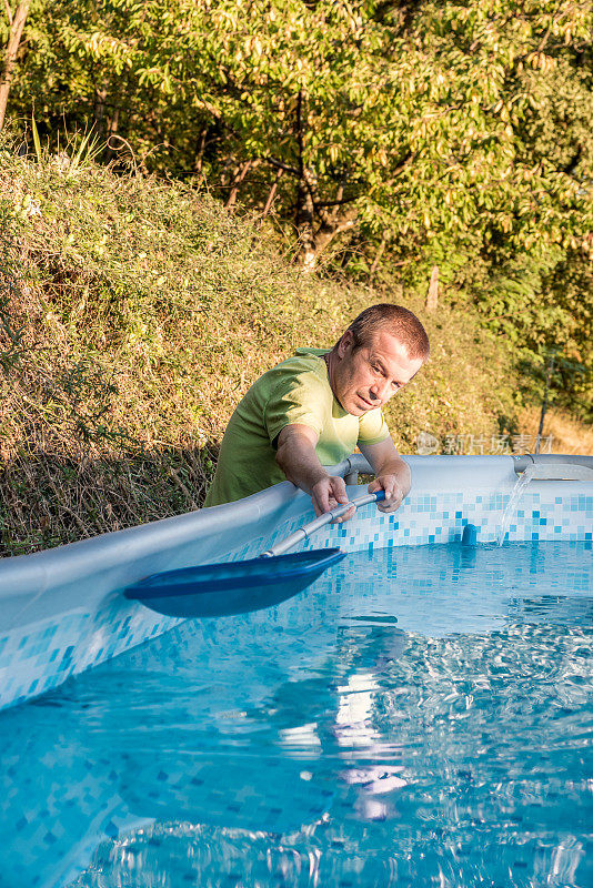 一名男子在后院清洁便携式游泳池，用网去除水中的叶子和昆虫