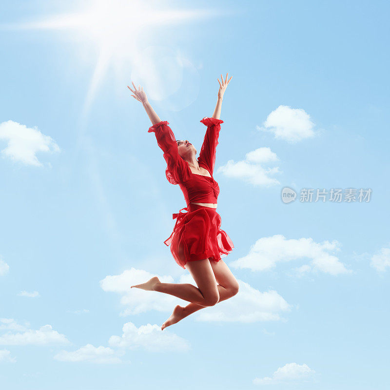身着红色连衣裙的年轻女子在空中跳跃，高举双臂，蓝天和太阳为背景