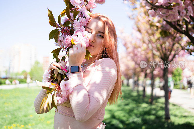 年轻美丽的时尚女孩在一个阳光明媚的日子里戴着帽子摆姿势附近盛开的树与粉红色的花的肖像。春天，女孩在一棵开花的樱花树旁。