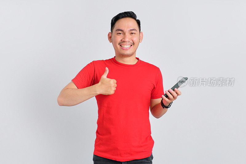 欢快的亚洲年轻男子在白色背景下使用手机，并显示竖起大拇指的手势