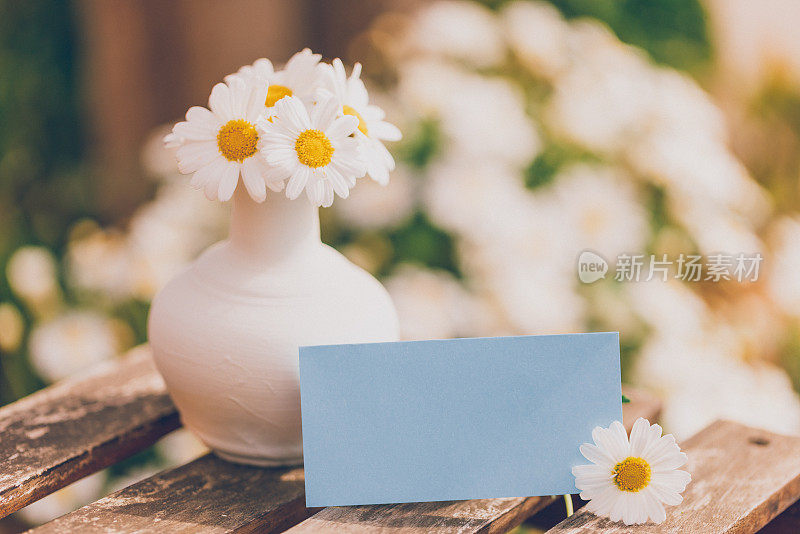 可爱的白色花瓶，上面插着白色雏菊，一张蓝色的空纸作为复活节、春夏短信的原型，放在一个小乡村桌子上