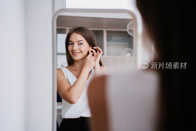 年轻迷人的意大利女子站在镜子上戴耳环露齿微笑准备约会。开朗的白人女孩在阳光明媚的早晨在家里。在酒店房间度假的漂亮模特。