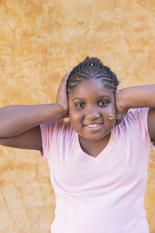 一个快乐的年轻美国黑人女孩的肖像，在户外墙的背景前微笑着捂着耳朵