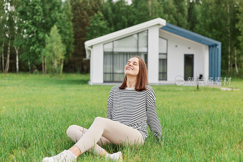 轻松愉快的女人坐在草地上，以现代房子为背景，在院子里休息，带着幸福的微笑仰望，享受阳光明媚的夏日。