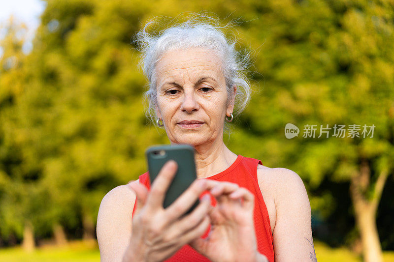 穿着红色运动服使用智能手机的资深拉丁女性。白发苍苍的老太太在公园里玩手机