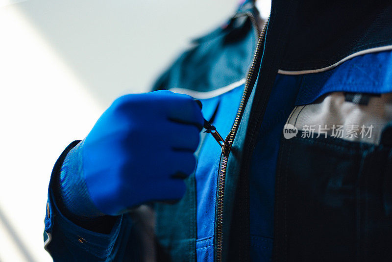 一个工人的手的特写，在工业仓库环境中，戴着安全手套拉上蓝色夹克的拉链。