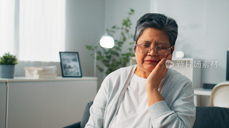 亚洲老人坐在家里的沙发上抚摸着牙齿问题的脸颊、因牙齿敏感而不开心的奶奶、牙齿疼痛的老年妇女