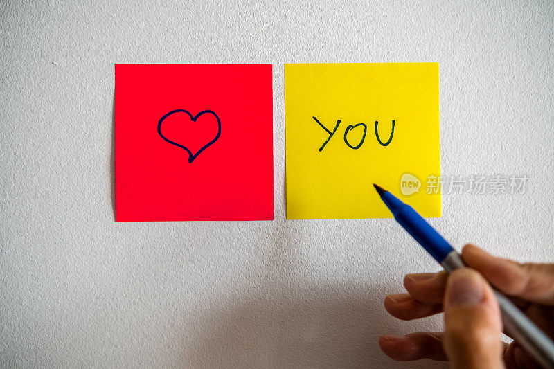 一个红色的柱子上画着一颗心，旁边是一个黄色的柱子，白色的墙上写着“你”，一只手拿着一支记号笔。观点。