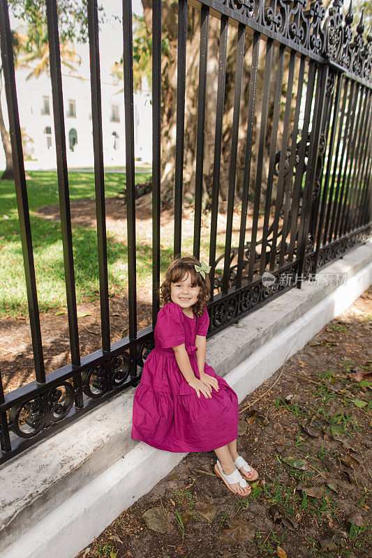 可爱的古巴裔美国三岁小女孩，棕色卷发，棕色眼睛和橄榄色肤色，穿着亮紫色连衣裙，蝴蝶结和白色凉鞋，站在佛罗里达州棕榈滩湖小径的黑色栏杆前
