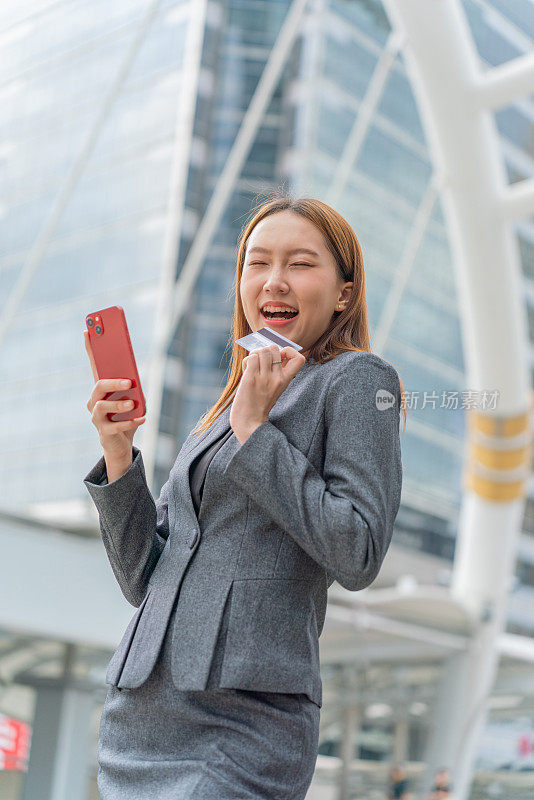 一名年轻的亚洲女商人穿着灰色的办公套装，在商业区用她的智能手机使用信用卡进行在线交易，她微笑着自信